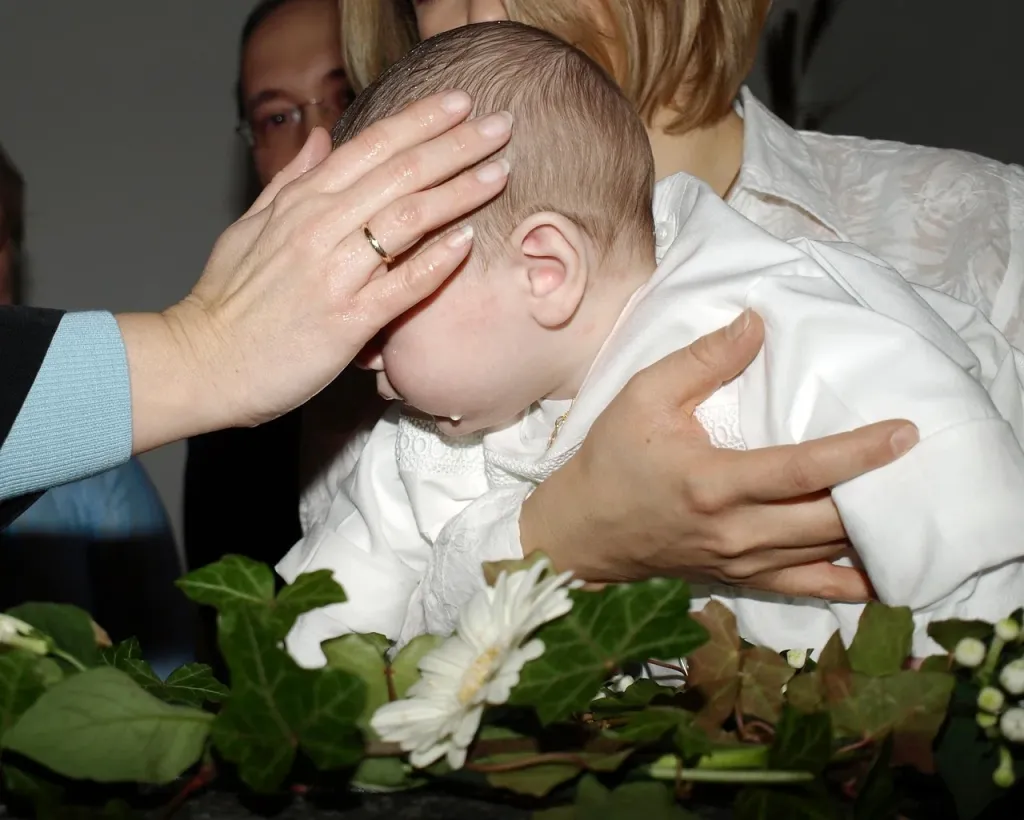 Elegancka biała sukienka do chrztu z koronki przewieszona przez drewniane krzesło w jasnym, przytulnym wnętrzu.