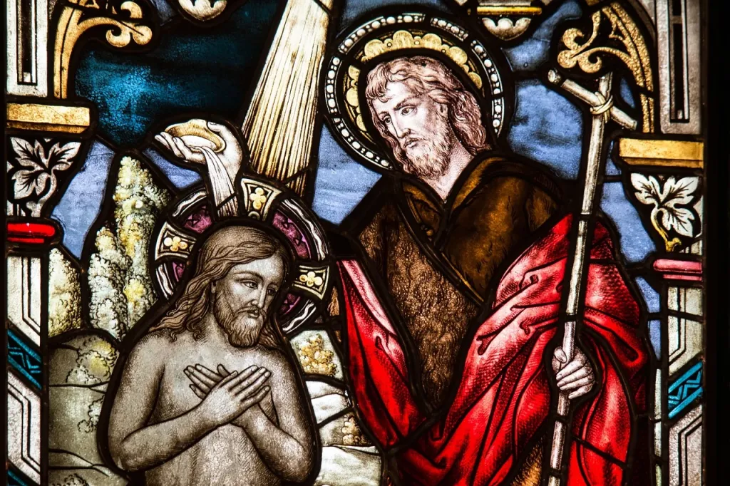 Srebrny krzyżyk i koronkowa czapeczka na drewnianym stole w ciepłym świetle świec, symbolizujące pamiątkę chrztu świętego.